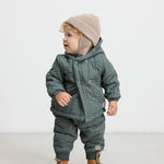 Baby iført støvet grønt termotøj og beige hue, står og kigger til siden