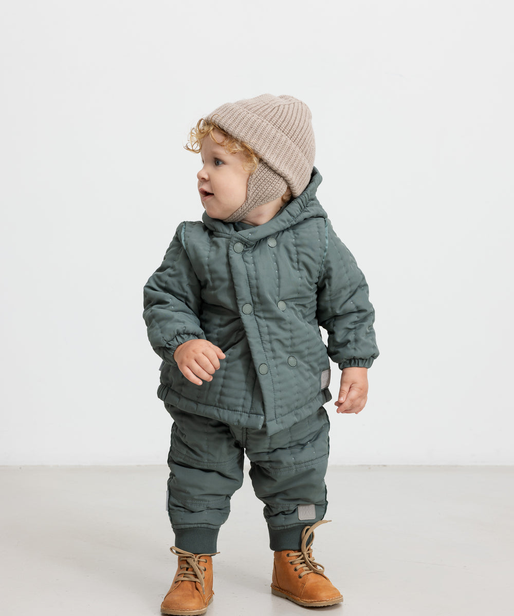 Baby iført beige strikhue og mørkegrøn heldragt, står og kigger til siden