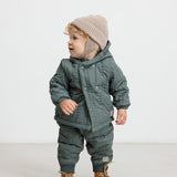 Baby iført beige strikhue og mørkegrøn heldragt, står og kigger til siden