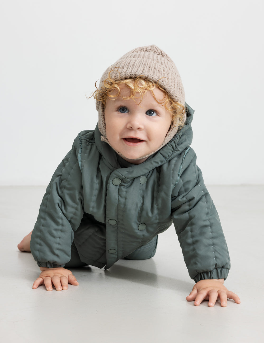 Baby iført støvet grøn termojakke og hue, kravler på gulvet.