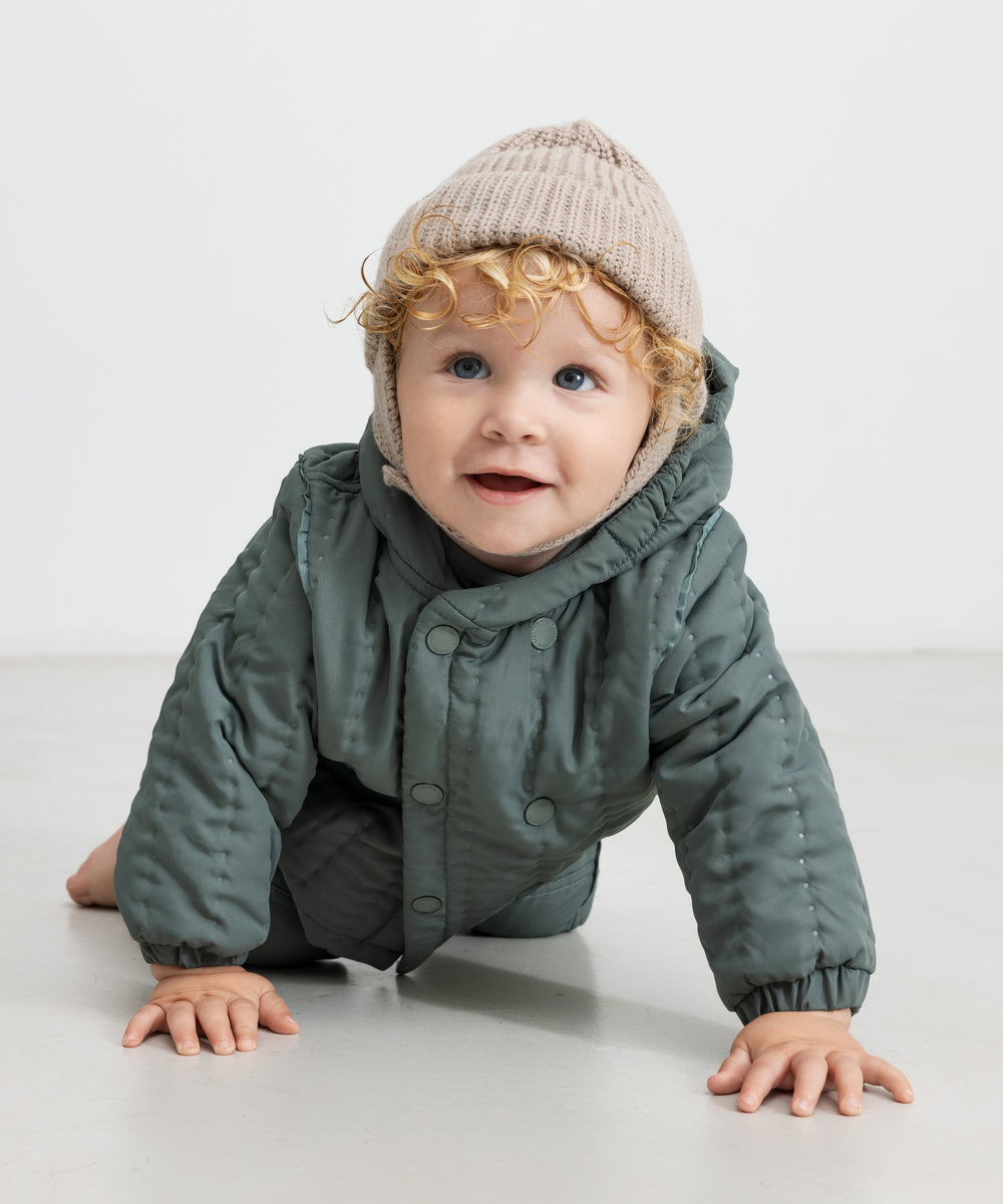 Baby iført beige strikhue og mørkegrøn heldragt, kravler mod kameraet