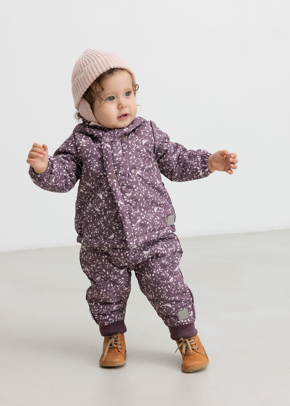 Baby iført lyserød strikhue og lilla heldragt, står og smiler