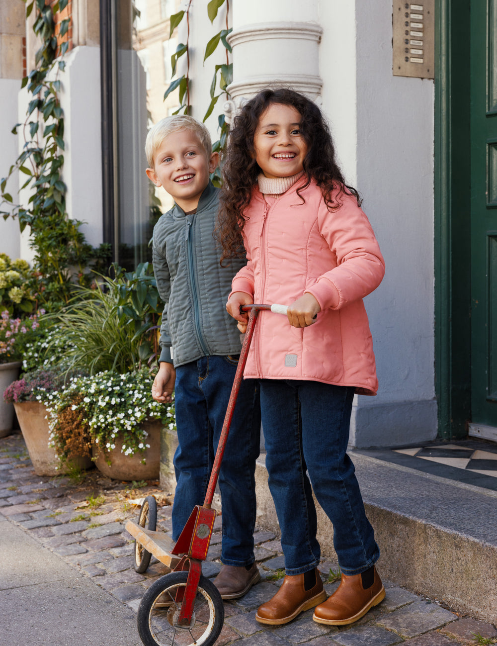 Børn foran en dør, dreng iført støvet grøn termojakke, pige i lyserød