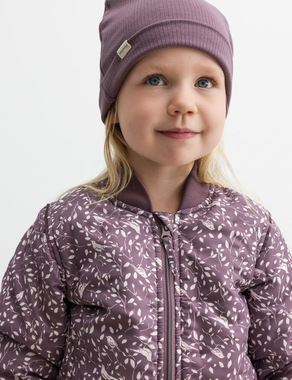 Barn iført lilla ribbeanie og jakke med print, smiler.