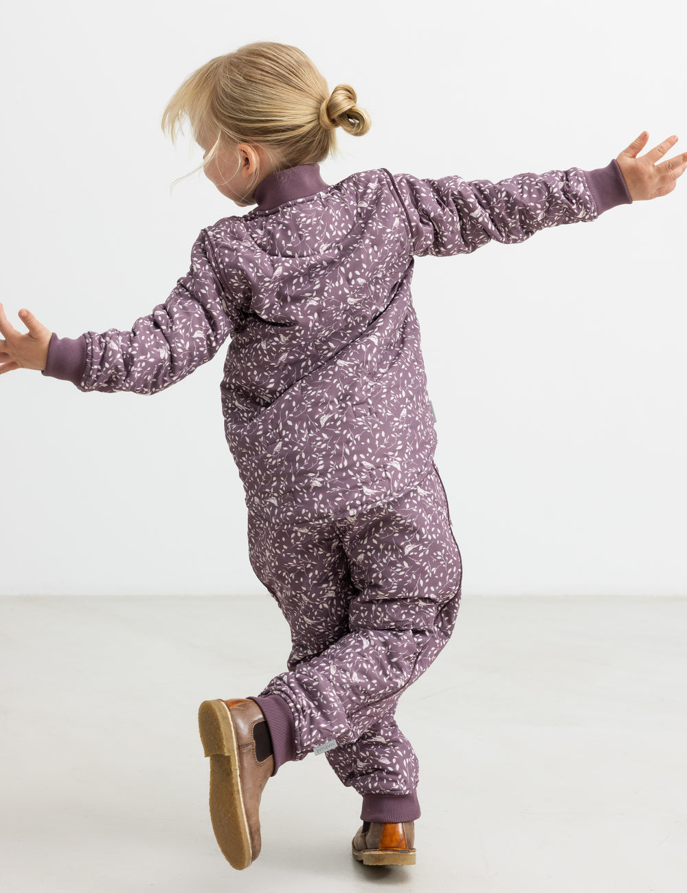 Barn iført støvlilla termobukser og jakke med print, hopper med armene ud.