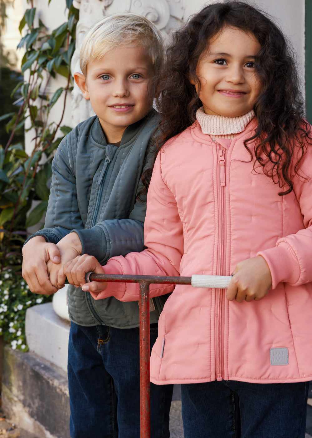 o børn holder en scooter, drengen iført støvet grøn termojakke og pigen lyserød