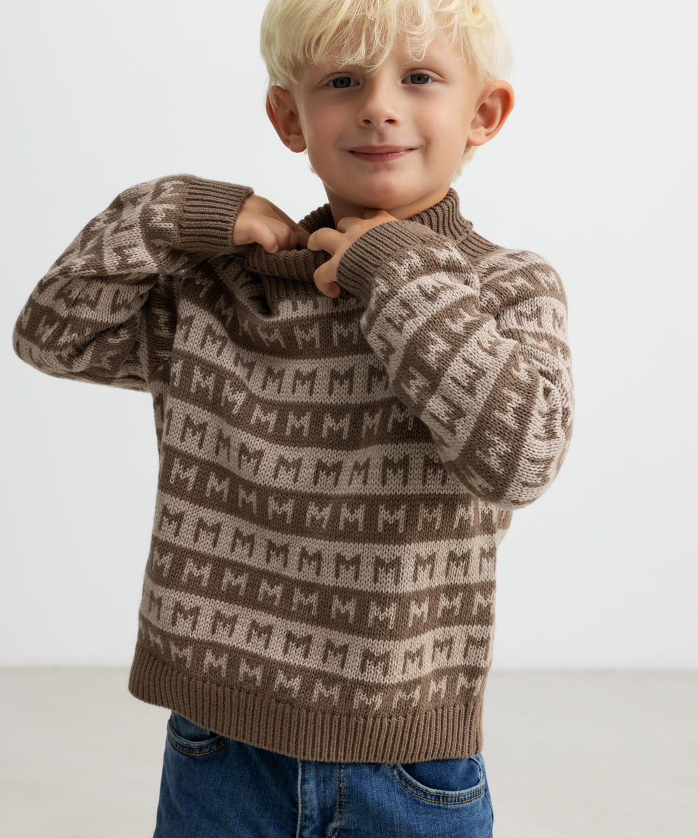 Barn tager en brun M-mønstret striktrøje på, smilende