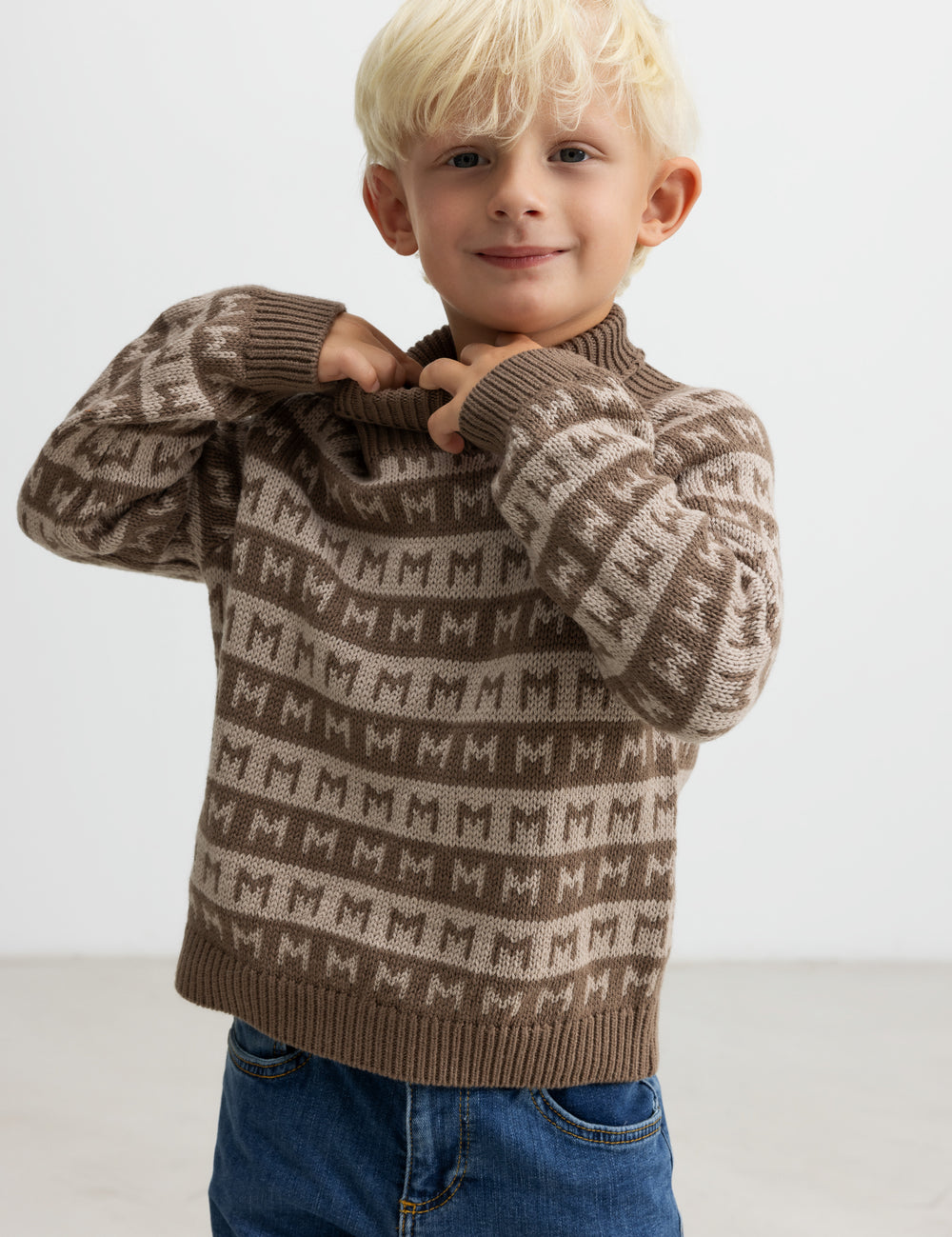 Barn tager en brun M-mønstret striktrøje på, smilende.