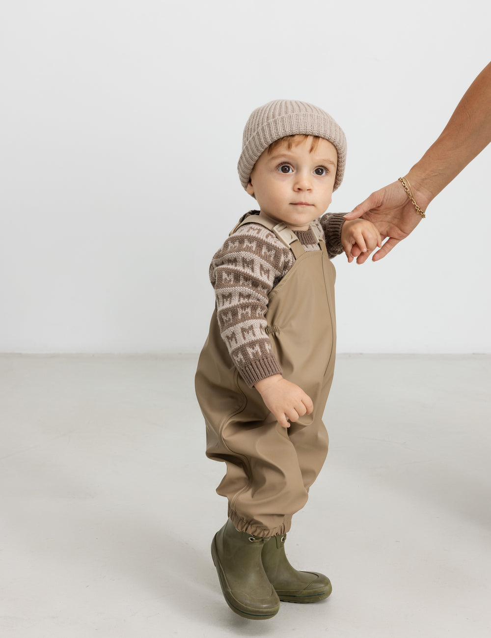 Baby iført brun M-mønstret striktrøje, termobukser og hue, holder en voksen i hånden.