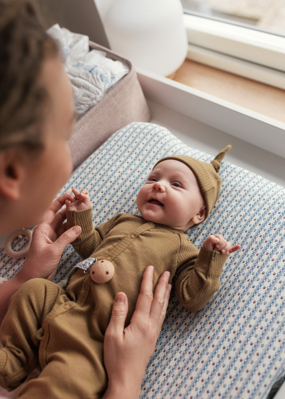 Mor leger med baby iført brun ribstrikket hue og heldragt ved puslebord.