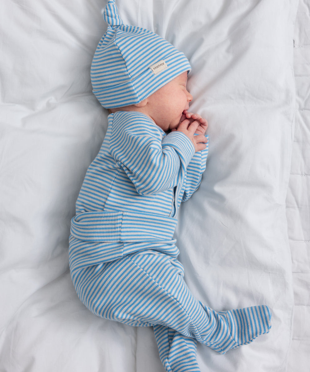 Baby iført blå- og hvidstribet body og matchende hue, sovende på hvidt tæppe