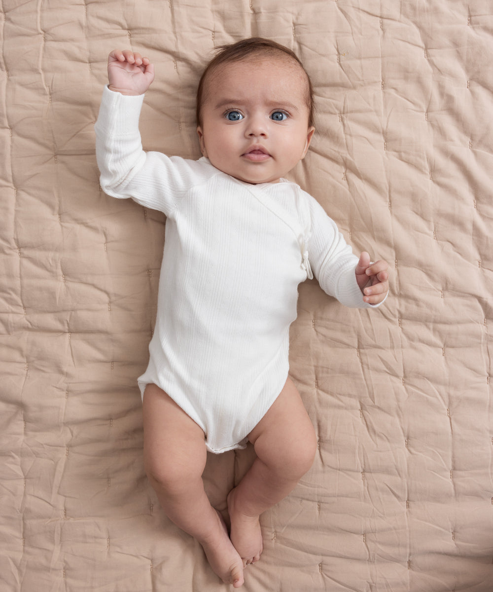 Baby iført hvid pointelle slå-om body liggende på quiltet tæppe