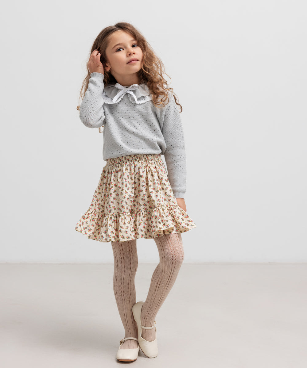 Pige iført grå strikbluse og nederdel, står og kigger væk