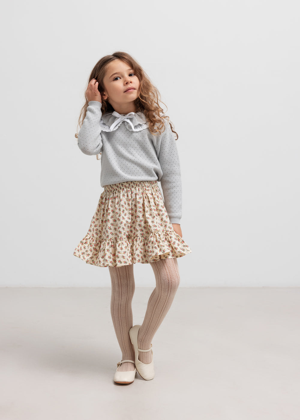 Pige iført grå strikbluse og nederdel, står og kigger væk