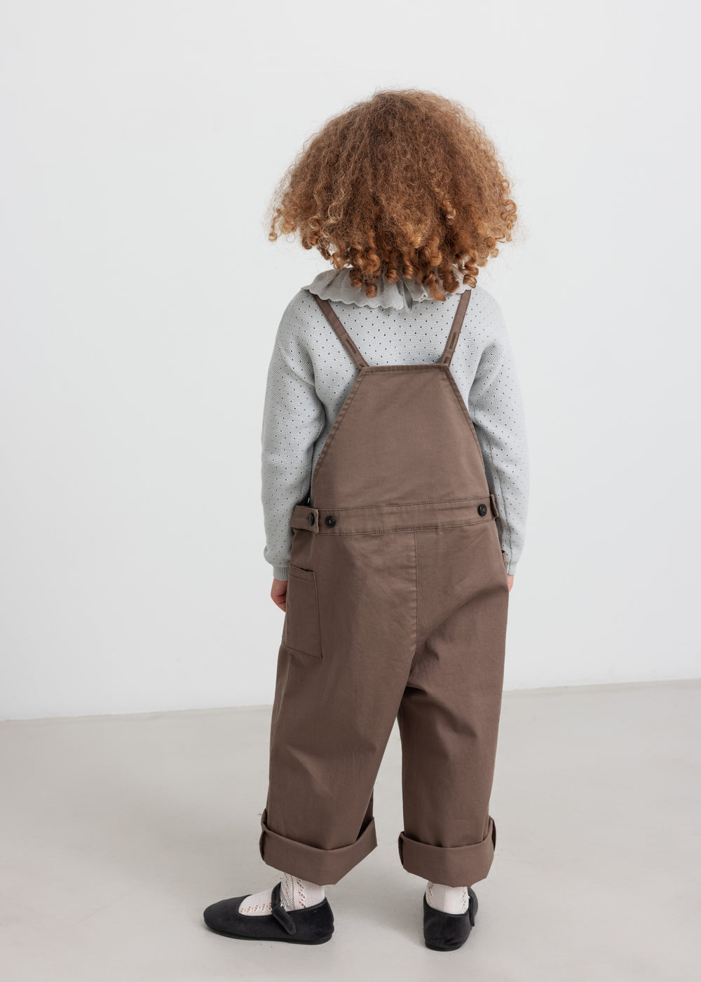 Pige iført brune selebukser og grå strikbluse set bagfra