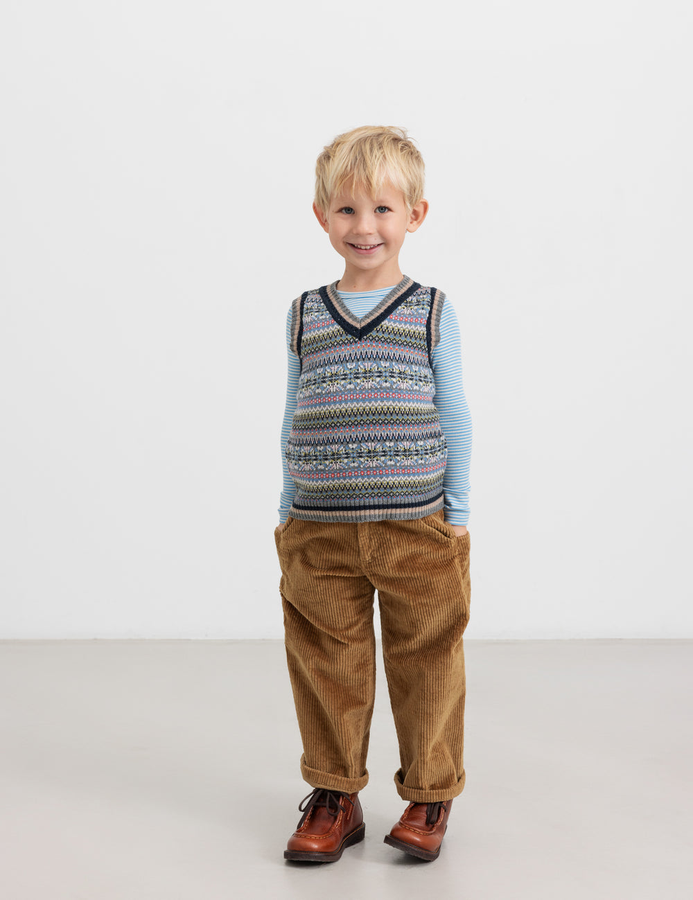 Dreng i brun buks og farverig strikvest med V-hals.