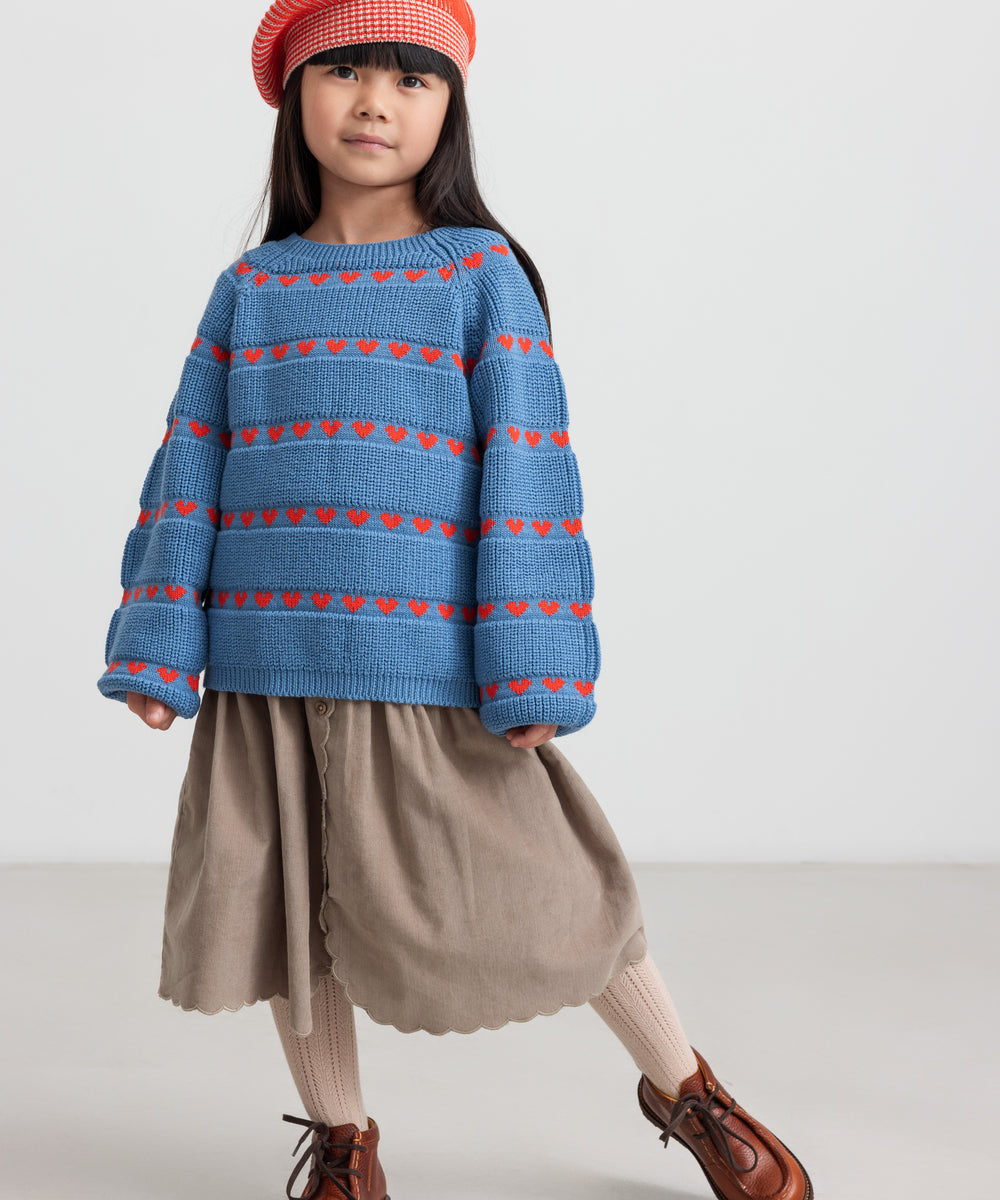 Børn iført blå striktrøje med røde hjerter, kombineret med brun nederdel og rød hue