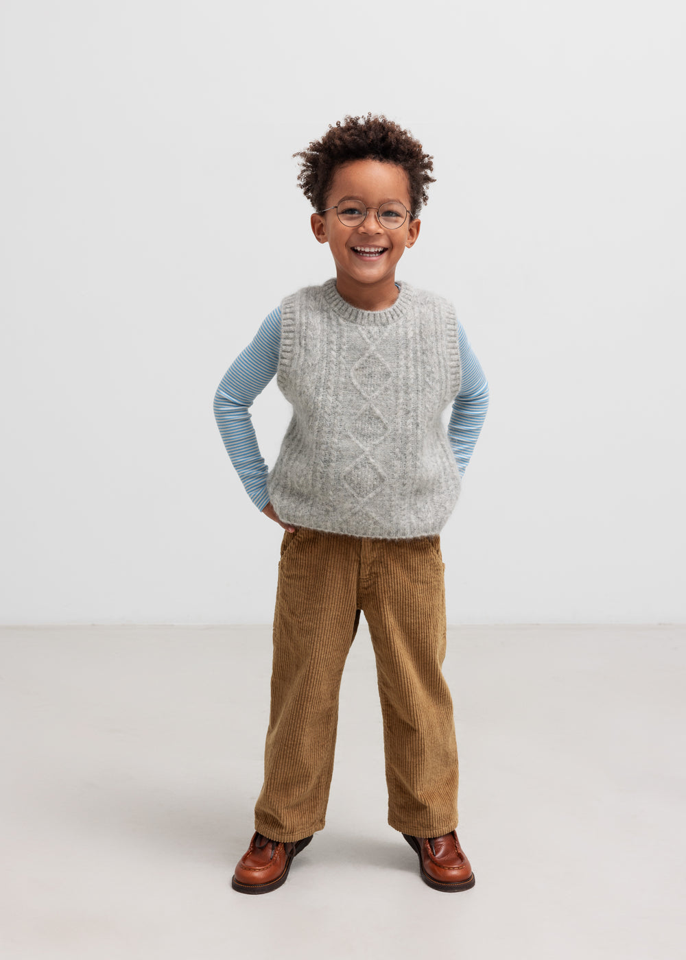Dreng iført blå og hvid stribet bluse med grå strikvest og brune bukser