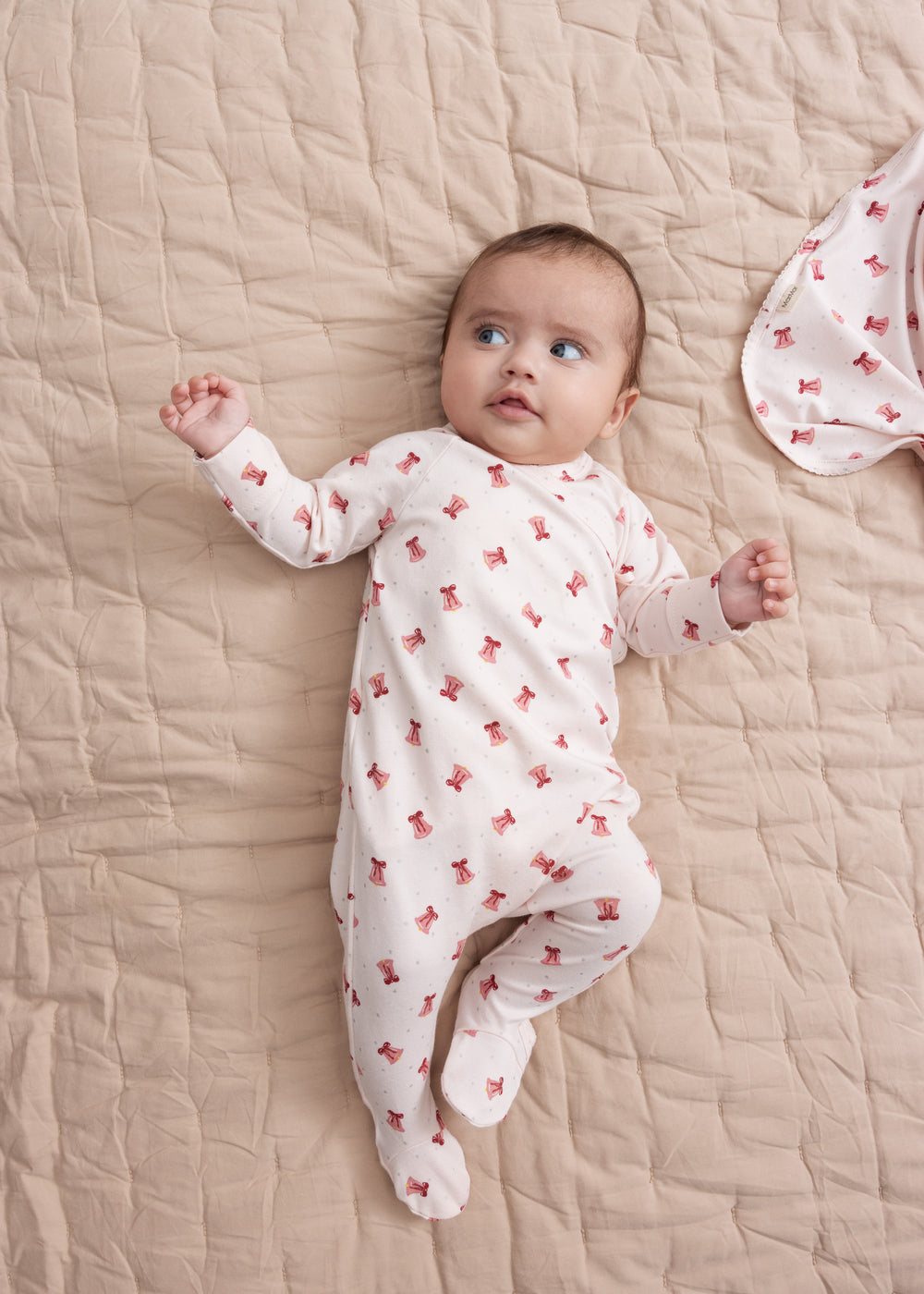 Baby iført cremefarvet heldragt med rødt juleklokkeprint liggende på tæppe