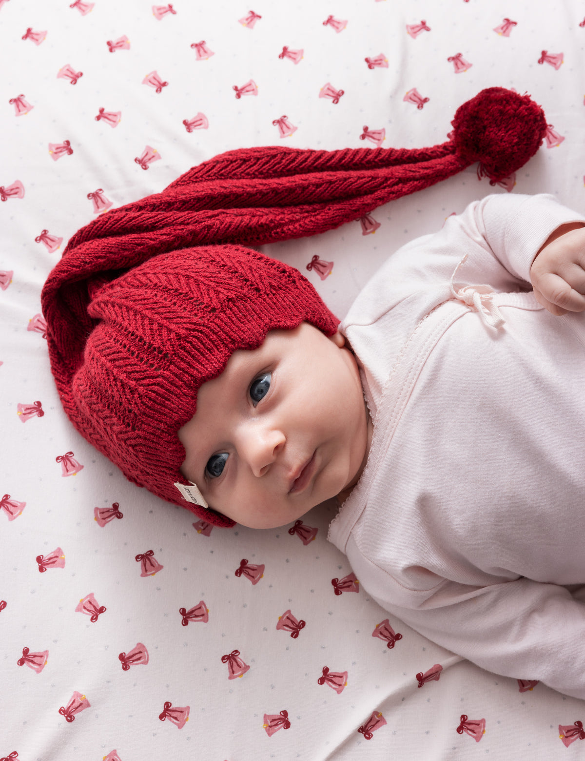 Nyfødt baby i lyserød body med rød nissehue liggende på lagen med julemønster