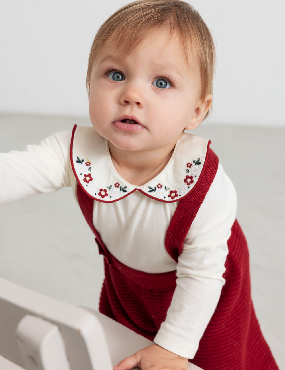 Baby wearing red knitted suspenders and white Christmas blouse with collar and floral embroidery
