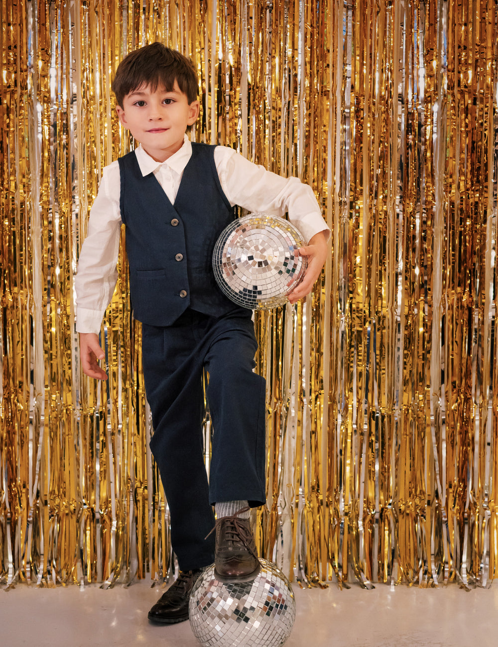 Dreng iført festtøj til børn mørkeblå bukser og hvid skjorte står foran gyldent festgardin med discokugle
