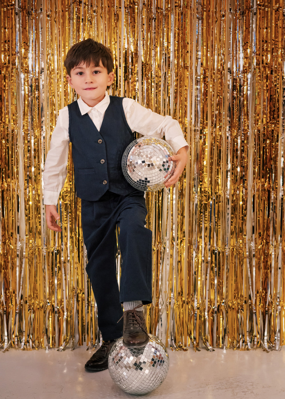 Dreng iført festtøj til børn mørkeblå bukser og hvid skjorte står foran gyldent festgardin med discokugle
