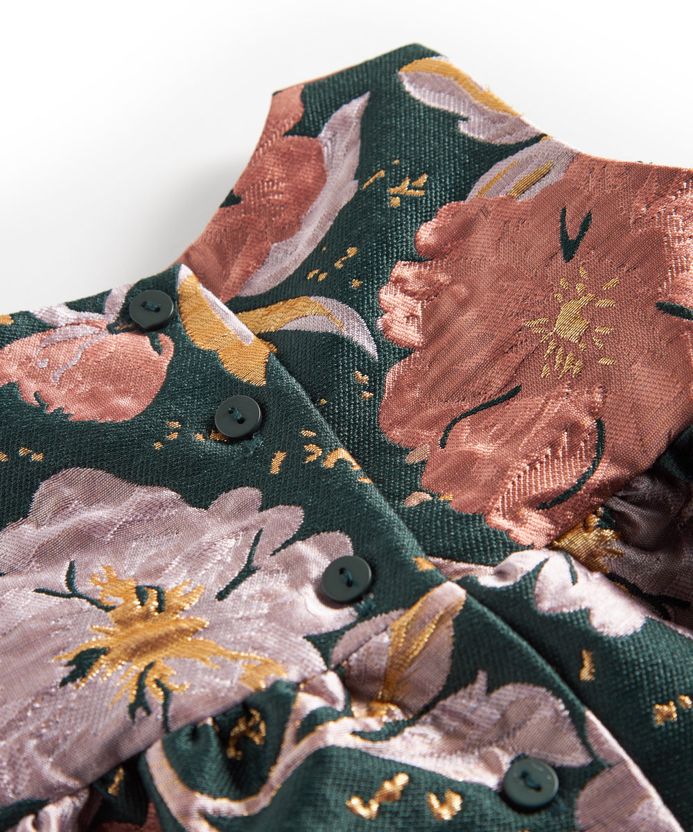 Nærbillede af mørkegrønt glansstof med blomsterprint og gyldne detaljer på festkjole til baby