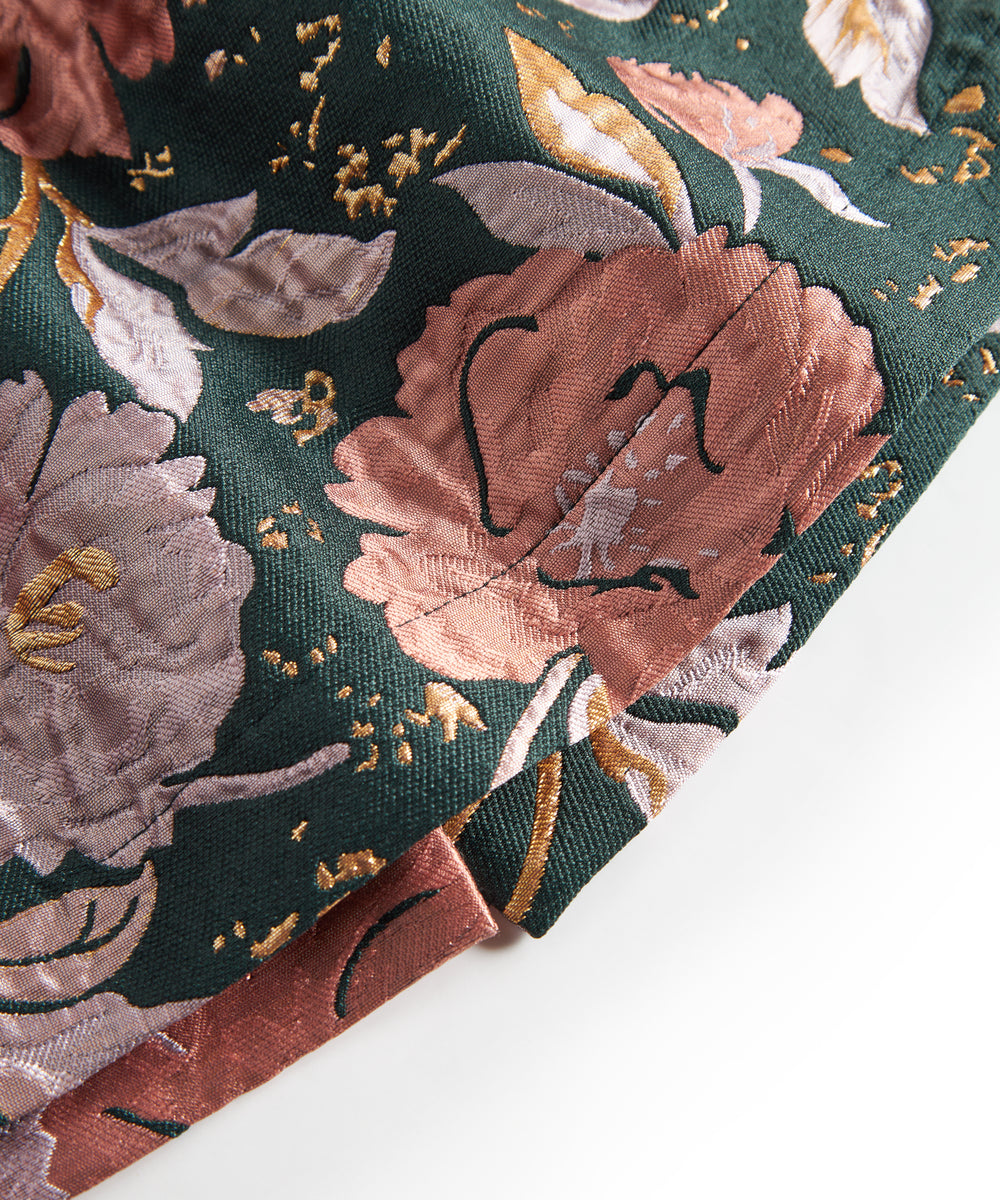 Nærbillede af mørkegrønt glansstof med blomsterprint og gyldne detaljer på festkjole til baby