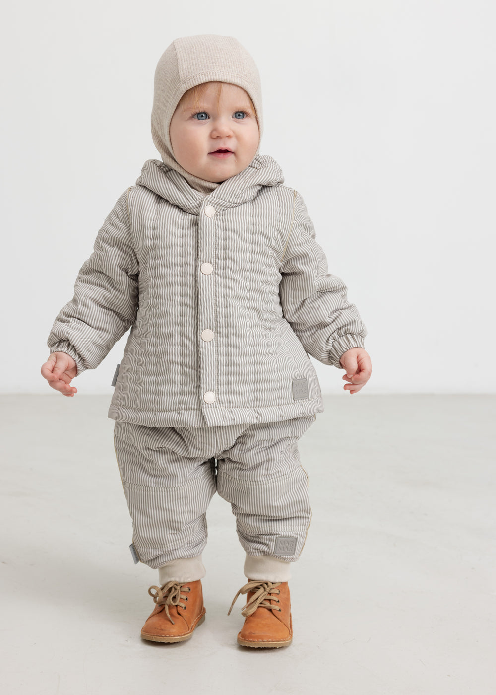 Baby wearing a plaid thermal jacket and pants with buttons, as well as a beige knitted hat.
