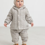 Baby wearing a plaid thermal jacket and pants with buttons, as well as a beige knitted hat.