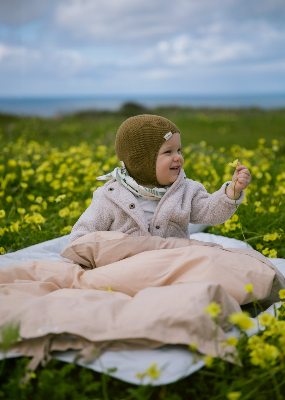 Baby wearing beige clothing and a brown knitted hat sitting on a blanket in the park.