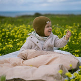 Baby wearing beige clothing and a brown knitted hat sitting on a blanket in the park.