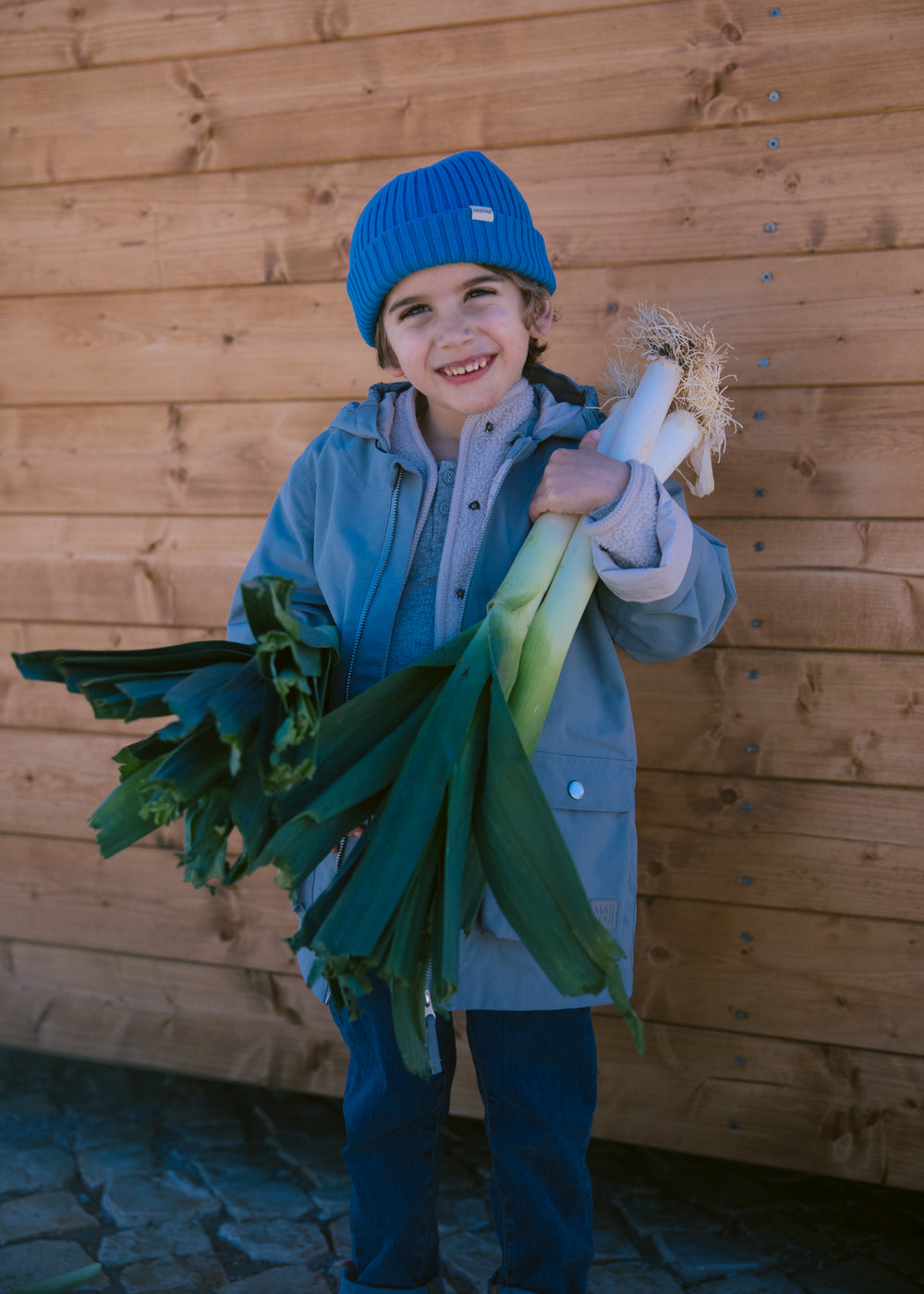 Child wearing a blue knitted hat and blue clothing with carrots in their hands.