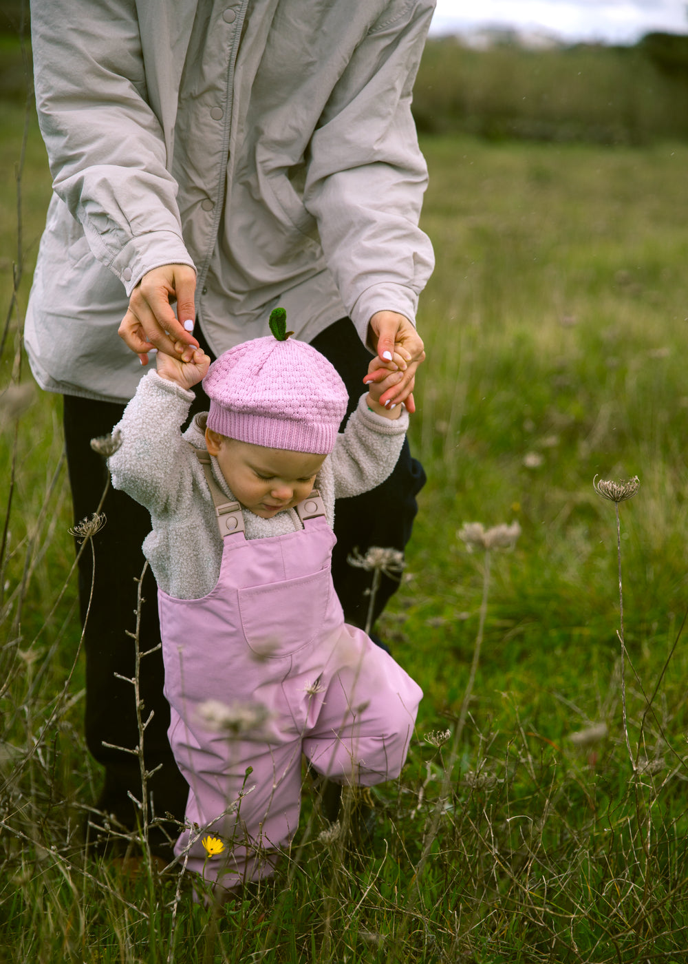 Baby wearing bright red overalls and a matching hat, walking supported by an adult.