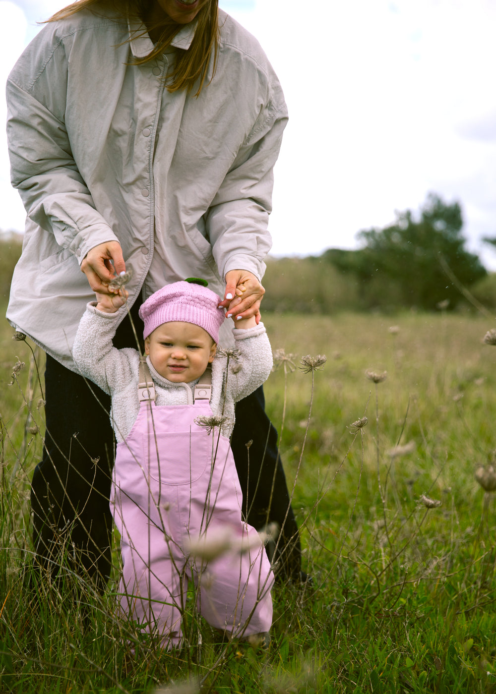 Baby wearing bright red overalls and a matching hat, walking supported by an adult.