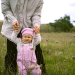 Baby wearing bright red overalls and a matching hat, walking supported by an adult.