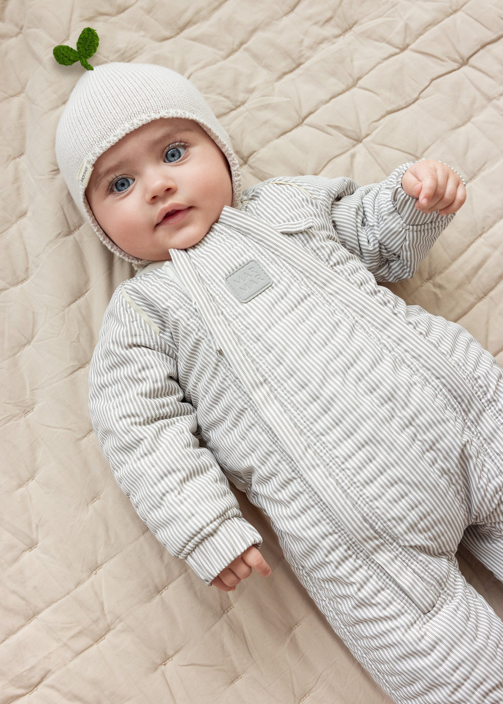 Baby wearing a striped thermal and a beige hat lying on a blanket.