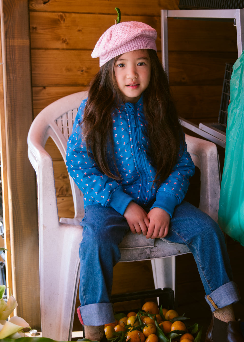 A child in a blue thermal jacket with a floral print and a bright red knitted hat sitting on a chair.