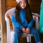 A child in a blue thermal jacket with a floral print and a bright red knitted hat sitting on a chair.