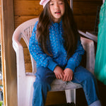 Child wearing a bright red knitted hat and a blue jacket sitting on a chair at the fruit stand.