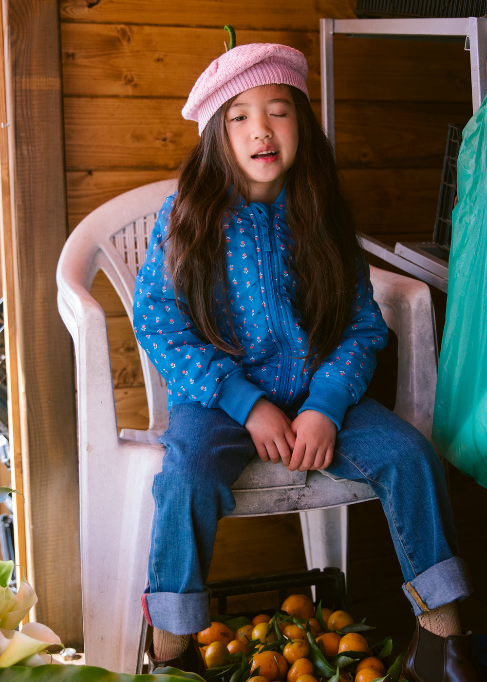 A child in a blue thermal jacket with a floral print and a bright red knitted hat sitting on a chair.