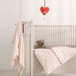 Baby swaddles in light color with small red hearts and decorative trim in a cradle.