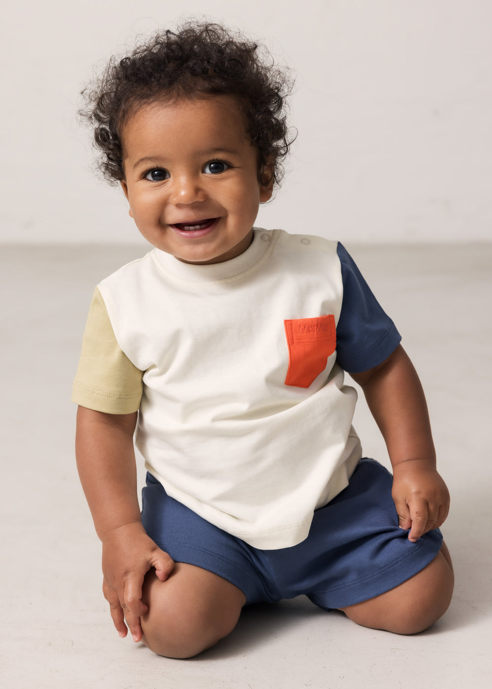 Baby wearing a raw white T-shirt with contrast sleeves and pocket.