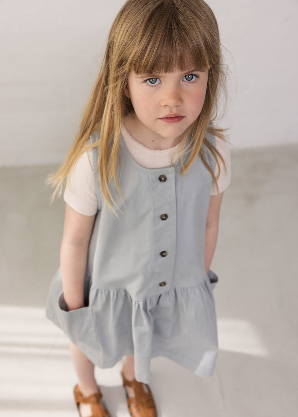 Child wearing a gray dress without sleeves with buttons and pockets.