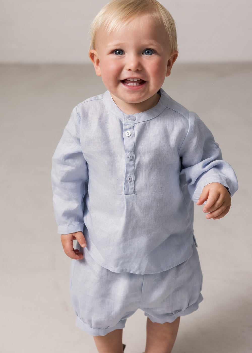 Baby wearing a long-sleeved light blue shirt with a crew neck and front buttoning, along with matching bloomers.