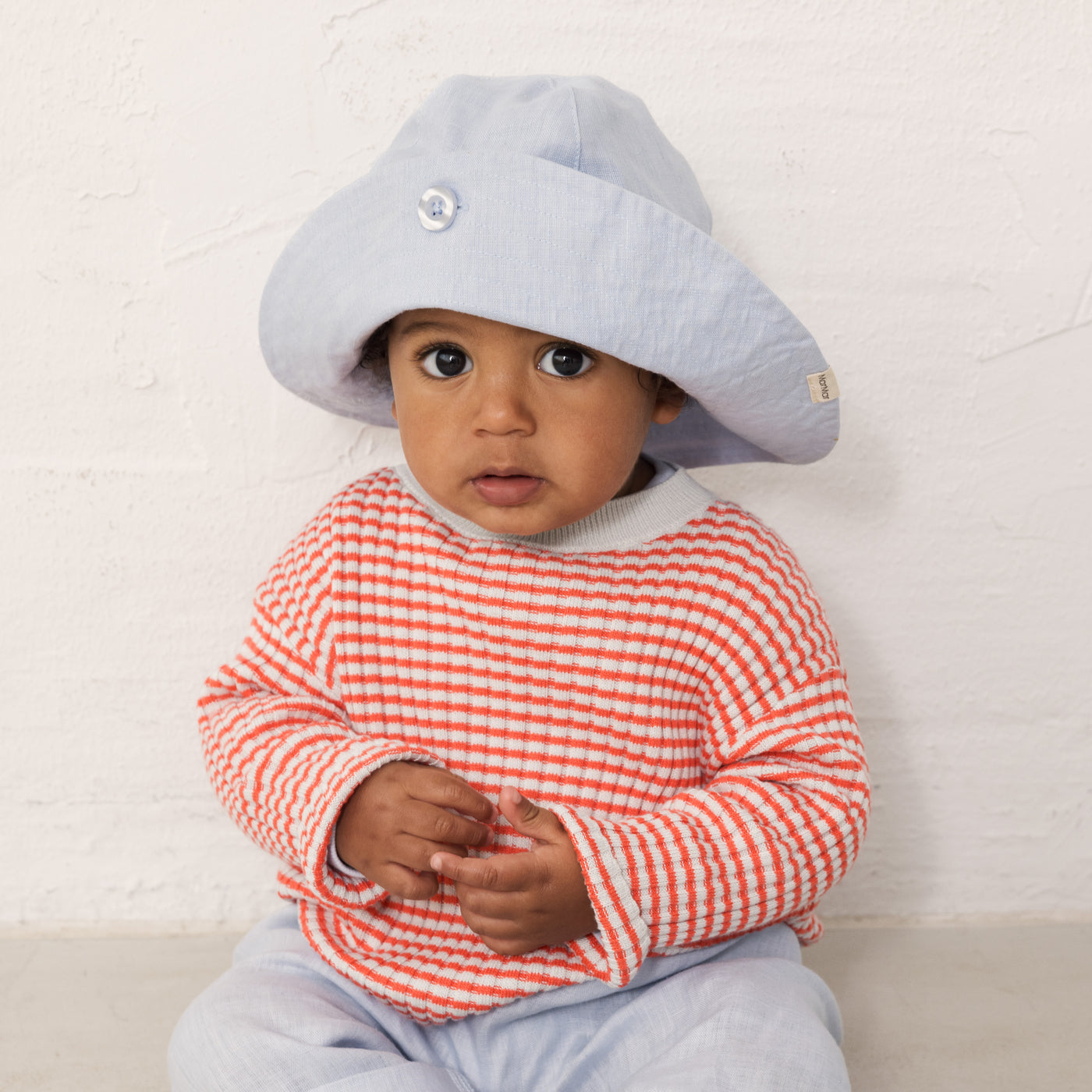 Baby iført lyseblå solhat, rød/blåstribet stik og lyseblå bukser