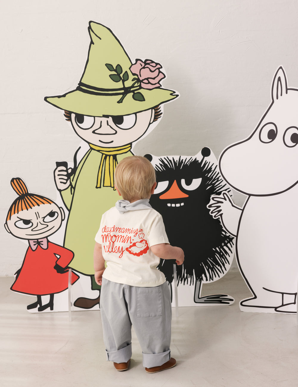 Baby wearing a white T-shirt with Mumin print and light pants together with Moomin figures.