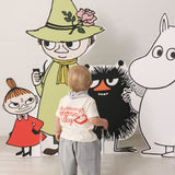 Baby wearing a white T-shirt with Mumin print and light pants together with Moomin figures.
