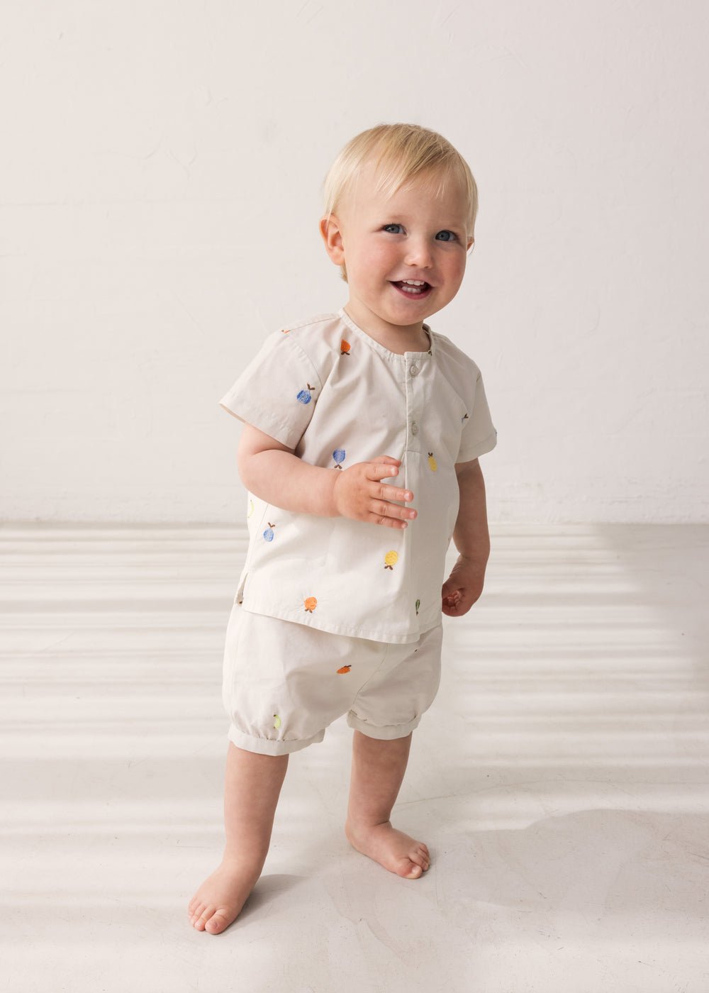Baby wearing light shorts with embroidered fruits and a matching top.
